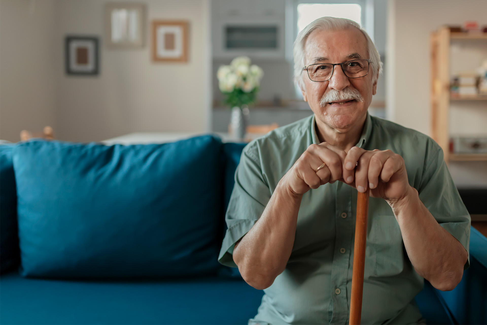 Älterer Herr mit Krückstock auf dem Sofa Lächelnder älterer Mann mit Brille und Krückstock sitzt auf einem blauen Sofa in einem gemütlichen Wohnzimmer.